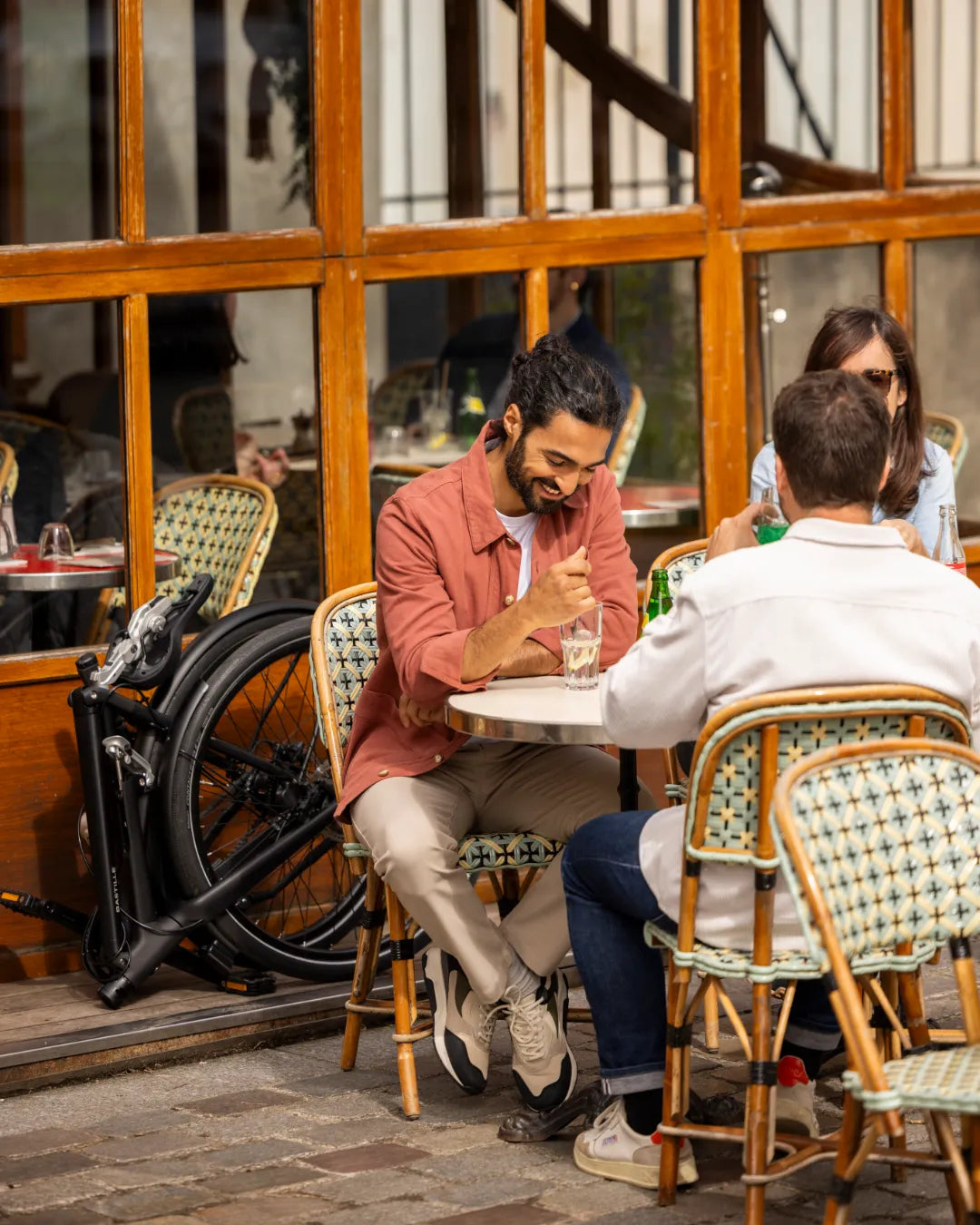 Un homme boit un verre en terrasse avec son vélo Bastille plié derrière lui.